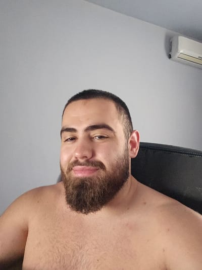 The image features a head-and-shoulders portrait of a young adult male positioned slightly off-center to the left. He has dark, short-cropped hair and a full, neatly trimmed brown beard. His brown eyes are directed towards the viewer, and he exhibits a subtle, closed-mouth smile. The subject is shirtless, revealing his bare chest and shoulders, which display visible body hair. A distinct mole is present on his upper left shoulder/arm area. The background consists of a plain, light grey wall. A dark, textured surface, appearing to be the back of a chair, is visible behind his right shoulder and ear. In the upper right quadrant of the image, a white wall-mounted air conditioning unit is partially visible against the grey wall.