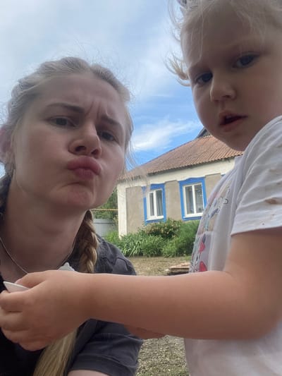 The image is a selfie taken outdoors, with two subjects visible: a young child and a woman. The child is in the foreground, looking directly at the camera with wide eyes and an open mouth. The woman is positioned below the child, also looking at the camera with a playful, puckered expression. Her hair is styled in a braid.

The background features a building with a tiled roof and windows with blue trim. Green foliage and grass are also visible, suggesting a rural or suburban setting. The sky above is blue with some clouds, indicating daylight.

The woman appears to be holding a white object in her hand, possibly a phone or a similar device, as she is taking the photo. The overall mood of the image is lighthearted and casual, likely a candid moment captured between the two individuals. No text is visible in the image.