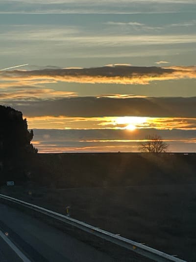 The image shows a vibrant sunset view from what appears to be a moving vehicle. The sky is filled with elongated clouds, illuminated by the setting sun in golden hues. These clouds create distinct vertical lines. To the right is a dark, undefined landscape with a bare tree. Part of the road and roadside barriers are visible at the bottom right, suggesting the photo was taken while driving. The perspective is somewhat skewed due to the angle of the shot.