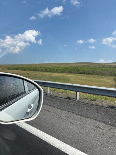 The image is a forward-facing view from a vehicle, looking out at a landscape. In the foreground, the side mirror of a white car is visible on the left, reflecting a hand holding a purple phone. The mirror is speckled with water droplets, suggesting recent rain or washing. The car's door handle is also visible. The road surface is asphalt with a white lane marking. A silver metal guardrail runs along the right side of the road. Beyond the guardrail, the landscape consists of rolling green hills covered in sparse vegetation, with a line of trees in the middle distance. The sky is blue with scattered white cumulus clouds. The scene is likely a roadside view in or near Chernogorsk, Russia, given the provided context of the city.
