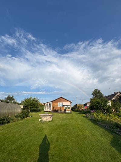 The image captures a rural scene in Sitno, Russia, with a vibrant rainbow arcing across a blue sky dotted with white clouds. In the foreground, a meticulously mowed green lawn leads up to a modest wooden house with a sunroom. To the left, a greenhouse or polytunnel stands partially visible. The house features a pinkish-brown exterior with white trim and windows. A stone well with glass jars sits on the lawn. In the background, other houses and trees are visible, contributing to the bucolic atmosphere. The shadow of a person holding a phone, presumably the photographer, is cast on the lawn. The overall impression is one of a peaceful summer day in the Russian countryside.