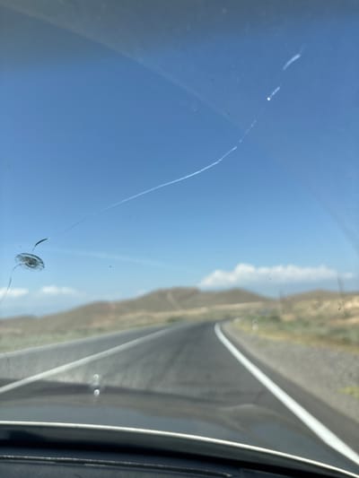 The image shows a close-up of a cracked windshield, viewed from inside a vehicle. The windshield is dirty with several visible smudges and specks. A prominent crack originates from a circular impact point and extends diagonally across the glass. The crack is long and thin, with some branching.

Reflected in the windshield is a bright blue sky with a few wispy clouds. The reflection also shows a landscape with hills and a road. The road appears to be paved and has a white line marking. The hills are dry and sparsely vegetated, suggesting a warm, possibly arid climate. The perspective of the reflection indicates that the vehicle is likely on a road in a rural or semi-rural area. The bright sky suggests it is daytime, likely mid-morning to late afternoon.

The overall impression is one of a journey interrupted by damage to the vehicle's windshield. There are no people visible in the image. The focus is on the crack and the reflections, which provide context for the setting. There is no visible text in the image.