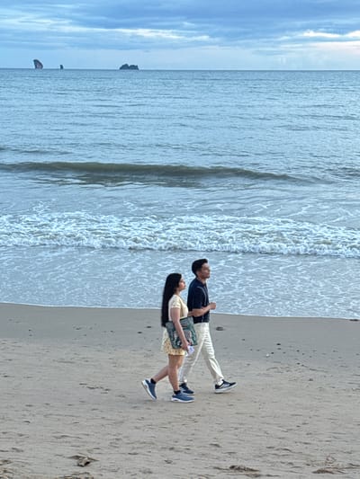 In Ban Klong Haeng, Thailand, a man and a woman walk along a sandy beach next to the ocean. The man, wearing a dark blue polo shirt and light-colored pants, walks to the right of the woman. The woman, in a short yellow dress and dark blue shoes, carries a patterned bag. The ocean has waves rolling onto the shore, and in the distance, several rocky islands are visible against a cloudy sky.