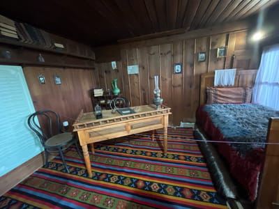 The image depicts an interior scene of what appears to be a historic room in the city of Zheravna, Bulgaria, likely a museum exhibit. The room is furnished in a rustic, traditional style with wooden paneled walls and ceiling.
In the foreground, a colorful striped rug with geometric patterns covers the wooden floor. To the left, a dark, curved-back chair with a woven seat sits next to a large, light-blue informational display.
Dominating the center of the image is a wooden desk with turned legs and decorative carved trim. On the desk, there is an oil lamp with a glass chimney, a black book or binder, and a dark bottle. To the left of the desk, a small stack of books rests on a shelf.
Behind the desk, a portion of a wooden bed is visible. It is covered with a dark red bedspread and a thick, shaggy blanket in shades of gray and blue. A decorative textile with a geometric pattern is draped across the foot of the bed.
The walls are adorned with several framed pictures, some appearing to be photographs. A green ceramic jug and a small glass are also visible on a shelf to the left of the desk, near a stack of books. The overall impression is of a preserved historical space, possibly a writer's or scholar's study.