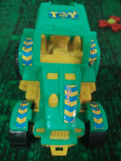 A green and yellow toy tractor labeled "Toy Farmer" sits on a green surface. The tractor's front and sides feature blue and yellow chevron patterns with "Farmer" printed vertically. It has a cab without doors and yellow interior details. Black tires are visible at the front and partially at the rear. A red and yellow object is partially visible in the upper right corner.