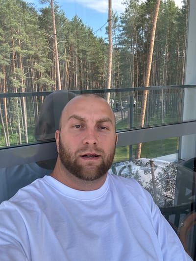 The image shows a man taking a selfie outdoors. He is bald with a beard and is wearing a white long-sleeved shirt. The man appears to be looking at the camera. The background is a reflection on a large window. Through the window, a dense forest of tall pine trees can be seen under a bright sky, suggesting it is daytime with fair weather. The reflection also shows part of the man's upper body and his white shirt. The scene has a casual feel. The location context is given as Мядзельскі сельскі Савет, Belarus, which implies a rural or semi-rural setting with significant forest coverage. There is no visible text in the image.