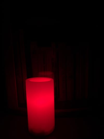 In Novaya Usman', Russia, a close-up, low-angle, full-frame shot captures a cylindrical, red LED candle set on a dark surface. The candle emits a vibrant, diffused red glow, illuminating its immediate surroundings. Behind the candle, a bookshelf filled with books is visible, though heavily obscured by darkness. The spines of some books bear faint, illegible text, suggesting they are in Russian. The overall atmosphere is dark and intimate, with the red light from the candle being the dominant source of illumination.