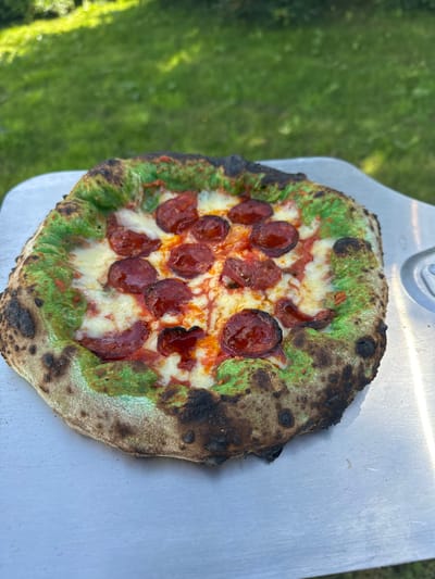 A pizza with a green-tinged crust and pepperoni is displayed on a metal pizza peel. The pizza is topped with melted white cheese and red sauce. The crust is charred in some areas. The background shows a blurry green lawn. The image was taken in the city of Moortown, Ireland.