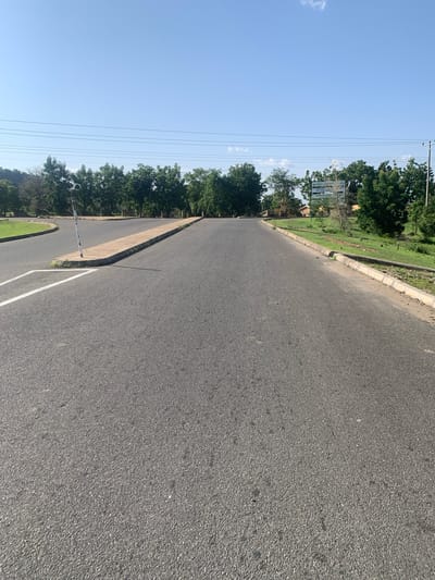 This image depicts an asphalt road in Boi A, Nigeria, under a clear, bright sky, suggesting it is daytime. The road curves gently to the left, bordered by a concrete curb and then a grassy area with scattered trees. In the distance, more trees line the landscape, and a few utility poles with wires are visible against the sky. A white signpost stands near the road, though the text on it is illegible. The scene appears calm and tranquil, with no people or vehicles present. The overall impression is that of a quiet, rural road on a sunny day.