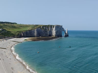 This media file depicts a panoramic view of the iconic white cliffs and arch of Étretat, France, on a clear, sunny day. The scene is set along the Normandy coast, where the rugged cliffs meet the turquoise waters of the English Channel.
The foreground features a pebble beach dotted with numerous people enjoying the summer weather. Some are sunbathing, others are strolling along the shore, and a few are venturing into the shallow waters where gentle waves break. Small boats and buoys are scattered across the sea, adding to the tranquil atmosphere of a recreational day.
Dominating the mid-ground and background are the majestic chalk cliffs, characterized by their steep faces and weathered formations. The most prominent feature is the natural arch, a striking geological formation that has become synonymous with Étretat. A smaller, spire-like rock formation stands sentinel nearby. The cliff tops are covered in lush green grass, with a golf course visible in the distance, hinting at the elevated landscape.
The clear blue sky and bright sunlight suggest it is midday. The overall mood is one of peaceful enjoyment of a beautiful natural landscape and seaside activities. No visible text is present in the media file.