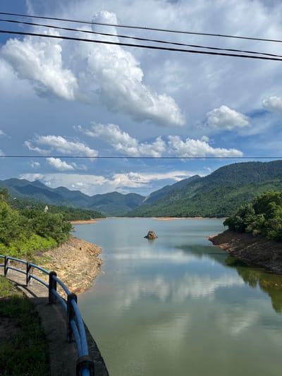 This is an outdoor, eye-level, vertical shot of a serene reservoir in Huế, Vietnam, with a mountain range in the background. The sky is partly cloudy with white cumulus clouds scattered across a bright blue expanse. Three black wires are strung across the top of the frame. The water of the reservoir is calm and reflects the sky and surrounding landscape. On the left bank, a rocky, dry embankment slopes down to the water, lined with a blue metal railing and a concrete path with some green grass. Further inland, lush green trees and foliage cover the lower slopes. In the distance, the mountains are covered in dense green forest. A small, rocky outcrop emerges from the water in the middle of the reservoir. The overall scene evokes a sense of tranquility and natural beauty.