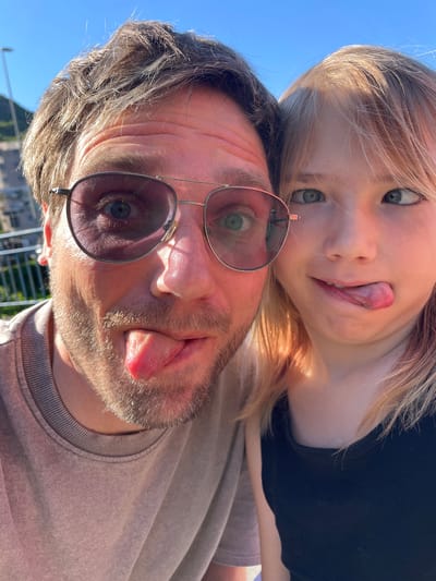 The image, rotated 90 degrees counter-clockwise from its likely upright orientation, captures a playful close-up of two individuals: an adult male and a child. Both are looking towards the camera with wide eyes and sticking out their tongues, indicating a lighthearted and fun moment.
The adult male, positioned in the foreground, has medium-length dark hair and a beard or stubble. He wears round-rimmed sunglasses, through which his light-colored eye is visible. He is dressed in a plain, light-colored (possibly grey or beige) crew-neck top. The child, positioned above and slightly behind the man, has long, blonde hair that is illuminated by the sunlight. She wears a dark top, and her light-colored eyes are prominent.
The background consists of a clear, bright blue sky, signifying an outdoor setting with sunny weather, likely during the late morning or afternoon. Faintly visible below the man are distant buildings, possibly mountains, and a railing, suggesting the photograph was taken from an elevated vantage point in Podličak, Montenegro. No visible text is present in the image.