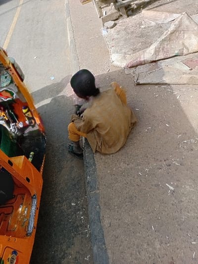 The scene depicts an outdoor street environment in Sharada, Nigeria, under daylight conditions. A single individual, appearing to be a child or young person, is seated on a concrete curb, observed from behind. This individual wears a light brown, long-sleeved tunic over similar-colored trousers and dark sandals. Their head is bowed, and their hands are engaged with an indiscernible object or activity on the ground directly in front of them at street level. To the left of the individual, an orange three-wheeled motorized vehicle, consistent with an auto-rickshaw, is parked on the asphalt street, parallel to the curb. The vehicle exhibits decorative elements and chrome accents. The street features a faded yellow lane marking. The ground area adjacent to the curb on the right, which appears to be a dirt or unpaved sidewalk, contains scattered white debris resembling wood shavings and a large, crumpled piece of light-colored material, possibly a tarp or bag.