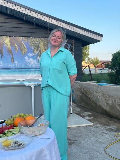The image captures a woman in a light blue suit standing next to a table laden with food outdoors in Новая Чернушка, Russia. On the table, covered with a white tablecloth, are platters containing fruits, vegetables, and salads. Behind the woman, a dark wooden building features a palm tree backdrop and string lights along its roofline. A concrete structure and a wire fence with vegetation can be seen in the background. The sky is clear and blue.