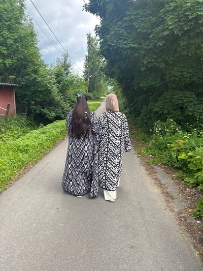 Two individuals, viewed from behind, walk along an asphalt path in Kommolovo, Russia. The person on the left has long, dark hair adorned with a blue butterfly clip. They are wearing a long, black and white patterned robe. The person on the right wears a light brown hijab and a matching black and white patterned robe over light-colored pants. The path is bordered by lush green vegetation and trees. In the background to the left, a small red building with a white fence is visible, along with power lines overhead. The sky is overcast.