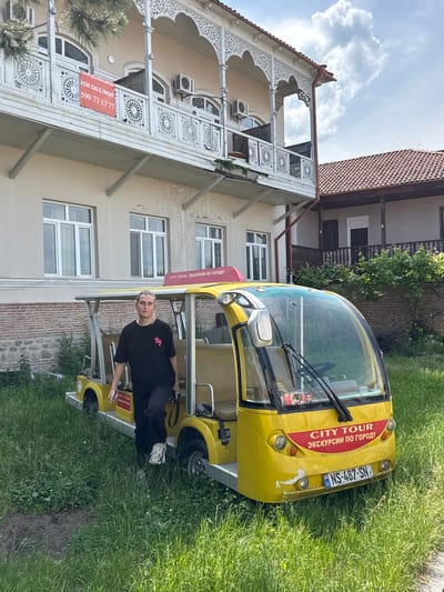 A young person with blonde hair, wearing a black t-shirt with a pink graphic and dark pants, stands next to a yellow, open-air tour vehicle in an outdoor setting. The vehicle has a red "CITY TOUR" banner above its windshield. Behind the vehicle is a light-colored building with a decorative balcony and a red-tiled roof. A red "FOR SALE/RENT" sign is visible on the balcony. The ground is covered in green grass, suggesting a park-like or garden environment. The scene appears to be in Sighnaghi, Georgia, a city known for its historic architecture and scenic views.