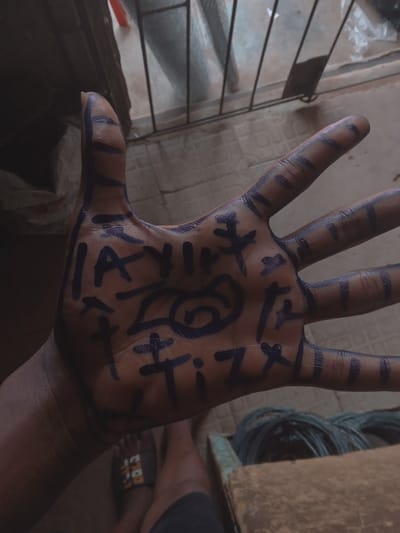 A photograph shows an open human hand with markings in a dark blue or black ink covering the palm and fingers. The markings appear to be stylized symbols or characters. The background is partially obscured, but a wire fence is visible, along with a blurred interior space. Part of a person's legs and feet wearing sandals are also visible. The image was taken indoors. There is no verifiable information about the city of Okpanam, Nigeria.