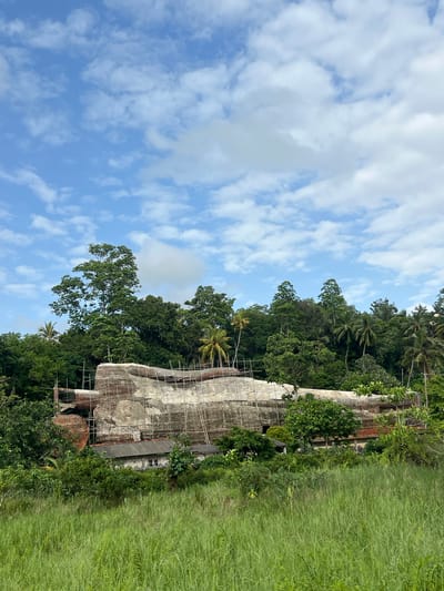 The image shows a building undergoing renovation in Walahanduwa, Sri Lanka. Scaffolding covers a large section of the structure, obscuring details of the facade. The building is surrounded by dense vegetation, including trees and tall grass. The sky is blue with scattered white clouds.