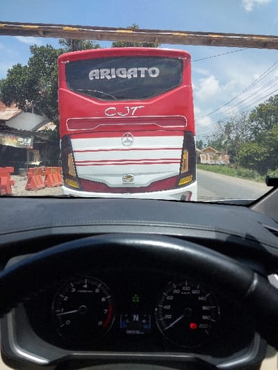 A forward-facing, eye-level photograph captures the rear of a red and white passenger bus on a paved road in Kulu, Indonesia, viewed from the interior of another vehicle. In the foreground, the upper portion of a black steering wheel and the vehicle's dashboard are visible. The instrument cluster displays a speedometer at 0 km/h, a tachometer at 0 RPM, the gear selector in Neutral ('N'), and an odometer reading 095918 km, with a green indicator light illuminated.
The bus, positioned directly ahead, features "ARIGATO" in white capital letters across its top rear window and "CJT" below it. Its lower rear section is white with red horizontal stripes, centered by a silver Mercedes-Benz logo. Yellow tail lights are visible at the bottom corners.
To the left of the bus, orange traffic barriers line the roadside, behind which a building with a reddish-brown roof and green foliage is visible. An overhead structure, appearing to be a wooden or metal beam, spans horizontally across the upper frame, above the bus. The road extends into the background on the right, bordered by trees, utility poles with power lines, and distant buildings, all under a clear, bright blue sky with scattered white clouds. The scene suggests a stationary position for the viewer's vehicle behind the bus during daylight hours.