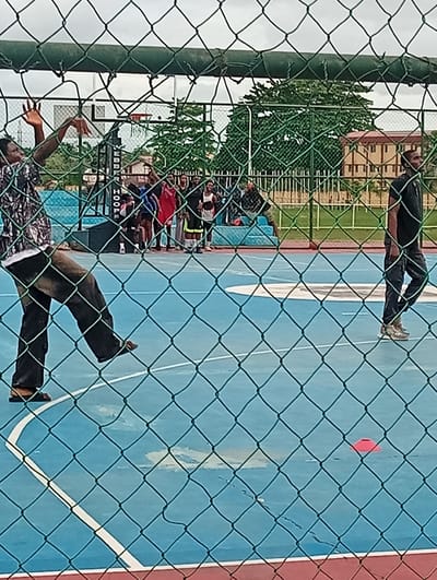 A daytime, eye-level outdoor shot taken from behind a green metal wire fence depicts a group of people on a blue basketball court, in Ijanikin, Nigeria. On the left of the frame, a man wearing a patterned, long-sleeved shirt and dark pants appears to be mid-motion, his left arm extended upwards. In the background, a group of people are clustered near a blue bench, observing the activity. To the right of the group, another man wearing a black shirt and dark pants is walking along the court. An orange cone sits on the court. A basketball hoop and the word "HOOPS" is visible in the background. Buildings and trees are visible in the background, under a cloudy sky.