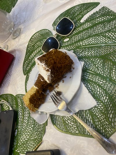 The image depicts a partially eaten slice of chocolate cake on a white plate. The cake is layered with white frosting and has a crumbly texture. There is a silver fork next to the cake on the plate, which is resting on a green leaf-shaped placemat. A pair of sunglasses is partially visible at the top of the image, resting on the placemat. The scene is set on a white tablecloth. The location is in Buea, Cameroon, based on the coordinates provided (latitude: 4.145, longitude: 9.23). A portion of a black cell phone is visible in the lower left corner of the image. A toothpick is visible in the lower right corner. The image appears to be taken from a slightly downward angle. The lighting is bright and there are no people present.