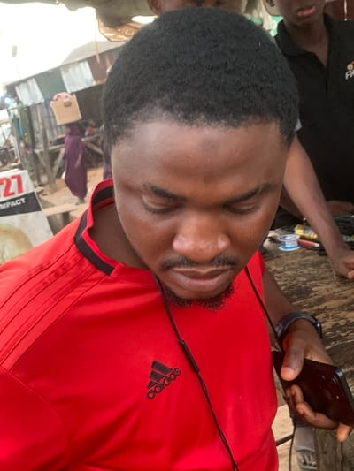 A man is angled, looking down at a black phone in his right hand. He's wearing a red Adidas shirt with black stripes and the Adidas logo. Black earphone wires are visible across his chest. His eyes are closed, and he has short dark hair and a beard. A wrist-worn device is visible on his left arm. People and structures form a busy outdoor background.
