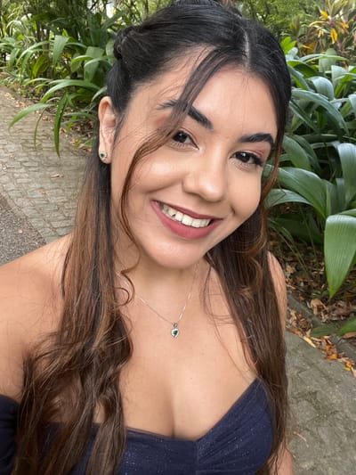 A female individual, approximately 20-30 years old, is depicted in a close-up, selfie-style photograph taken in the city of Camaragibe, Brazil. She has medium-brown hair with visible waves, styled partly pulled back and partly framing her face. Her complexion is medium, and she is wearing facial makeup, including defined eyebrows, eyeshadow, and a matte rose-toned lipstick. She is adorned with small stud earrings, each featuring a green central gemstone surrounded by clear stones, and a silver-toned necklace displaying a heart-shaped pendant with a green gemstone centerpiece also encircled by clear stones. Her attire consists of a dark, possibly navy or black, strapless garment, appearing to be a dress or top, made of a subtly textured or glittery fabric. The individual is actively smiling broadly towards the camera, revealing both upper and lower teeth. The background, out of focus, indicates an outdoor setting characterized by lush green foliage and a visible paved path or ground covering composed of irregular stone pavers, suggesting a park or garden environment under natural daylight.