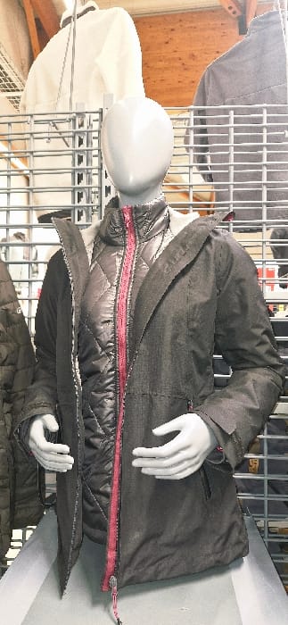 This image captures a headless, light grey mannequin displaying layered outdoor apparel within a retail store in Vendin-le-Vieil, France. The mannequin is positioned facing slightly right of center. It is dressed in a dark grey/black quilted inner jacket featuring a prominent, unzipped red central zipper. Over this, an open, dark grey/charcoal outer jacket with a hood is worn, revealing pockets with red zipper pulls near the waist and red accents on the cuffs. The mannequin's featureless head area and open hands are visible. In the background, metal grid display racks hold various items of clothing, including a white top and a grey jacket. Wooden ceiling beams are visible above the racks, and the mannequin stands on a light-colored display platform.