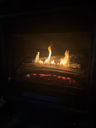 The image depicts the interior of an active fireplace in Roquiague, France. Within the dark, enclosed firebox, a fire is burning. Multiple yellow and orange flames rise dynamically from a horizontal arrangement of partially consumed logs situated in the mid-ground. Beneath the logs, a vibrant bed of glowing red and orange embers mixes with ash, occupying the lower section of the fireplace. The internal walls of the fireplace appear dark, with the rear wall exhibiting a textured, possibly brick-like surface, and the overall structure is indicative of metal or stone construction. No individuals or external objects are visible in the scene.
