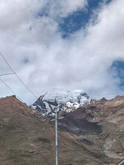The image shows a snow-capped mountain peak towering over rocky, barren hills. The mountain summit is partially obscured by clouds. In the foreground, a utility pole with wires stretches across the frame, obscuring parts of the mountainous landscape. The sky is a mix of blue and white clouds. The scene appears to be located in a remote, mountainous region, possibly in or around Padum, India, given the context. The overall impression is one of rugged natural beauty, with man-made infrastructure intruding on the landscape. There are no people visible in the image.