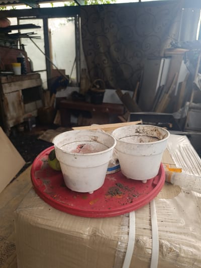 The image depicts an outdoor scene in Ostrogorka, Russia, focusing on two white plastic plant pots resting on a red plastic tray. The pots are dirty and contain what appears to be soil and some pinkish material, possibly decaying organic matter. One pot has a greenish object next to it, resembling a tennis ball. The tray itself is also dusty and speckled with soil.
The pots and tray are positioned on top of a large, rectangular object wrapped in clear plastic sheeting and secured with white strapping. The background suggests a cluttered, possibly abandoned or storage area, with various wooden planks, shelves, and what looks like a curtain with a swirling pattern. Sunlight filters through, casting shadows and highlighting the dust and debris in the environment. The overall impression is one of neglect and disuse. There are no people visible in the image.