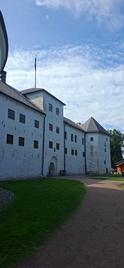 The image shows an exterior view of Turku Castle in Turku, Finland, on a partly cloudy day. The castle is a large, white stone structure with a dark-tiled roof. It features multiple windows with dark frames, some of which are visible in the wall. A flag flies from a pole on top of one of the towers. In the foreground, there is a well-maintained lawn and a gravel pathway. To the right of the castle, a small wooden building with a red roof is partially visible, along with a few distant figures of people. The scene conveys a sense of historical architecture and parkland.