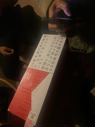 A person is holding a smartphone pointed at a pack of cigarettes in a poorly lit environment. The cigarette pack is red and white, and has Japanese text on it. The person's skin appears to be dark. A portion of another person's clothing is visible in the upper-left. The location appears to be indoors. There is no explicit information in the image to confirm it is located in the city of Montreuil, France.