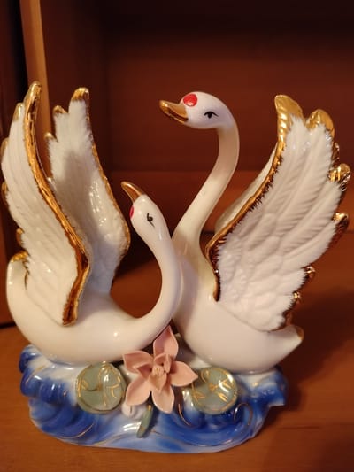 A porcelain figurine of two white swans with gold trim on their wings and beaks sits on a blue and white base decorated with gold accents. One swan's head is tilted towards the other, and both have red dots on their heads and painted black eyes. The base also features a pink flower and a light green lily pad with gold designs. The figurine is placed on a wooden surface, likely a table or shelf, in an indoor setting. The background is a dark brown, out-of-focus surface, possibly a wall or furniture. This item is not in Chișcăreni, Moldova.