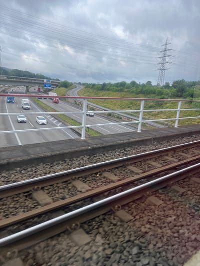 The image shows a view from a train in Bochum, Germany, looking out over a highway. The highway is multi-lane and curved, with several cars and trucks visible. There is a large power line tower in the background. The sky is overcast. The foreground of the image shows train tracks and ballast.