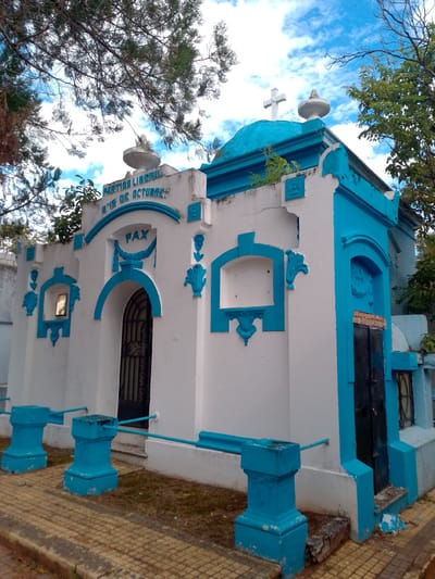 The image shows a mausoleum painted white with blue trim, located outdoors. Inscription reads "Partido Liberal 5 de Octubre PAX". A wrought iron gate provides access. A white cross adorns the top. The structure is flanked by trees and a partly cloudy sky is visible. The tomb is located in Asunción, Paraguay.