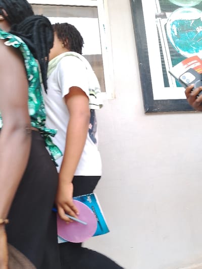 The image shows two young females in Ogboko, Nigeria. One is wearing a green patterned top and black pants. The other is wearing a white t-shirt and black pants, holding a blue book with a pink circular object on top and a pen. A cell phone and a picture frame are partially visible. They are next to a white wall and a window.