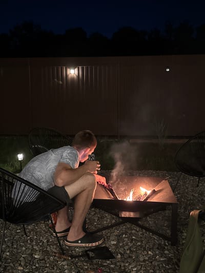The image depicts a young male sitting in a black wire chair next to a metal fire pit in an outdoor setting at dusk. He is wearing a grey and white patterned t-shirt, dark shorts, and black Crocs. The fire pit contains burning logs, producing smoke and flames. The ground is covered with gravel. In the background, there is a dark brown corrugated metal fence with two small solar-powered lights. Behind the fence, silhouetted trees are visible against a dark blue sky. The overall scene suggests a backyard or patio area.