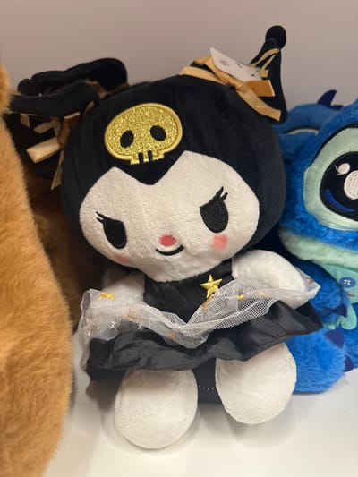 This media file is a close-up, overhead shot of several stuffed toys. The primary subject is a black and white plush toy with a witch-like hat and costume, featuring a gold skull emblem on its head. The toy has large, embroidered eyes and pink blush marks on its cheeks. It is adorned with black fabric, sheer white netting with gold stars, and gold ribbon accents.

Behind this toy, a portion of a blue plush toy, possibly a character like Stitch, is visible, with its distinctive blue fur and large eyes. Partially obscuring the bottom of the frame is a tan, furry object, which appears to be another stuffed toy or perhaps the fur of a pet.

The toys are arranged on a plain, light-colored surface, likely a shelf or table, creating a simple background that highlights the subjects. There is no indication of the time of day or weather. The setting appears to be indoors, in a domestic or retail environment where stuffed toys are displayed or stored. The overall impression is one of a collection of whimsical or themed plush toys. No discernible text is visible in the image.