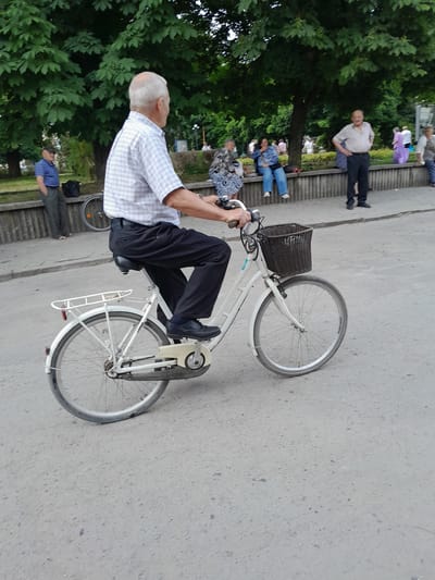 An elderly Caucasian man with gray hair, wearing a light-colored checkered button-down shirt, black pants, and black shoes, is riding a white bicycle with a black basket on the front. The man is positioned in the foreground, with the lower half of his body and the bicycle filling the center of the frame. The bicycle has a white frame, silver spokes, and a rear rack. The man's hands are on the handlebars, and he appears to be pedaling.

In the background, several other people are visible, mostly gathered on a concrete wall or sitting on benches under large trees. To the left, a man in a blue and white striped shirt and dark pants stands near a black bag. Further back, people are seen in various poses, some seated and conversing, others standing. The environment appears to be a park or public square, with mature trees providing shade and a paved surface underfoot. The overall lighting suggests an overcast day. The location is identified as Berezhany, Ukraine, implying an outdoor setting within the city.