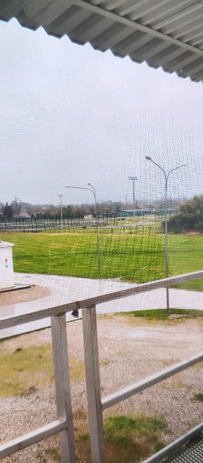 The image shows an outdoor scene, likely a sports field or park area, under a cloudy sky. In the foreground, a metal railing with vertical supports runs horizontally across the frame, suggesting a viewing platform or walkway. Beyond the railing, a large expanse of green grass covers the ground, with some patches of lighter gravel or dirt. A small white building with some red and blue markings is visible on the left side of the frame. In the middle ground, a fence with vertical posts separates the grassy area from what appears to be another section of the field or a pathway. Several tall light poles with multiple lamps are scattered across the landscape, indicating the area is equipped for nighttime use. In the background, a line of trees forms a distant border, with some structures and possibly more light poles visible amongst them. The overall impression is of an open, somewhat utilitarian outdoor space on a somewhat overcast day. There is no visible text in the image.
