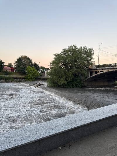 The image shows a moderately turbulent river flowing over a man-made weir. The river water is murky brown. On the left bank, there are buildings and trees. On the right bank, a large tree stands near a concrete bridge that spans the river. The sky is a clear, pale blue, suggesting it is either early morning or late evening. The foreground features a granite ledge and walkway.