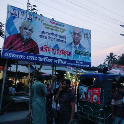 This medium-shot image depicts a lively street scene in Bangladesh during dusk or early evening, dominated by a large banner suspended above a bustling market. The sky in the upper half is a pale blue, crisscrossed by numerous power lines, with some palm trees visible in the distance.
The prominent banner extends across the upper frame, displaying festive Eid greetings in Bengali. It features two large portraits: a man with a shaved head and goatee in a red shirt on the left, and a man in a suit and tie on the right, identified as "মোঃ রাইসুল আলম" (Md. Raisul Alam), Member Secretary of the Bhola District BNP (Bangladesh Nationalist Party). Three smaller portraits are also visible at the top. The banner includes religious phrases like "বিসমিল্লাহির রাহমানির রাহিম" (In the name of Allah, the Most Gracious, the Most Merciful) and the patriotic slogan "বাংলাদেশ জিন্দাবাদ" (Long live Bangladesh). The text "ভোলাবাসীকে জানাই পবিত্র ঈদুল আযহার ঈদ শুভেচ্ছা" translates to "Wishing the people of Bhola a blessed Eid-ul-Adha." The banner is sponsored by "মোঃ জসিম শরীফ" (Md. Jasim Sharif), associated with a Bhola Sadar BNP committee.
Below the banner, a busy market unfolds with numerous people moving between makeshift stalls, many featuring corrugated iron roofs. The lighting is dim, indicating evening, with artificial lights illuminating the stalls and their wares. Several individuals are visible, including a man in the foreground talking on a mobile phone. On the right, a partially visible auto-rickshaw displays an advertisement featuring a woman and beverage products, with the text "KOPI" and "নিউ মুক্ত" (new release/new freedom). The scene conveys an everyday market environment, likely with a celebratory atmosphere due to the Eid greetings.