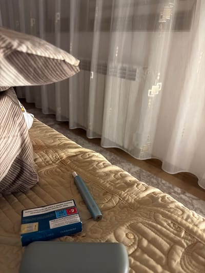 An indoor scene, likely a bedroom, is captured from a low angle. In the foreground, a light brown, quilted blanket covers a bed. On the blanket, to the left of center, is a partially visible pack of turquoise cigarette-like cartridges, with Russian text on the packaging. Next to the cartridge pack, to the right, lies a slender, light blue electronic smoking device with a metallic end. A striped pillow is visible in the upper left corner of the image. In the background, sheer white curtains hang, obscuring a window and a radiator. The overall lighting suggests an evening or dimly lit indoor environment. The image does not provide information about the city of Zmeitsa, Bulgaria.