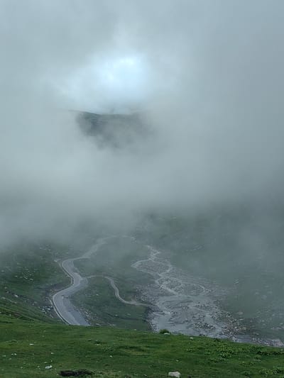 The image depicts a winding mountain road and a rocky riverbed nestled within a valley, enveloped by thick fog. The landscape is characterized by green, grassy slopes dotted with scattered rocks. In the upper portion of the image, a brighter area suggests the presence of the sky or sunlight obscured by the dense fog, with a distant mountain peak faintly visible through the haze. The road appears to be paved and shows signs of wear. The scene evokes a sense of isolation and the grandeur of nature in a remote mountainous region. The location is Marhi, India.