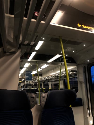 The inside of a train car is depicted at an angle. Blue, patterned seats are on the right with grey armrests. Yellow handrails run horizontally throughout the car. A sign on the wall contains train information. Overhead lights illuminate the train's interior. A digital sign displays text in yellow. Metal bars and a glass barrier are visible on the left. Outside, the sky can be seen through a window.