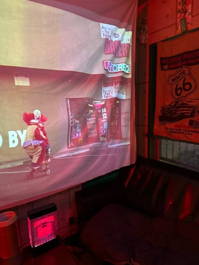 This image depicts a dimly lit room with red lighting. The scene features a projection on a large white sheet, showing a street with neon signs and a clown figure on a tricycle. The clown is wearing a red and yellow costume with white face paint. The projection also displays text, including "BY", "Sala", "SMIRNOFF", "GU TE Pan Bakery", and "BAYCRY ROBEO".
Behind the projection, there are several objects. A brown burlap banner with a "Route 66" logo and an illustration of a classic car is hanging on the wall. Below this, there's a glimpse of a dark brown leather couch. To the right of the couch, a Smirnoff vodka box is visible, illuminated by red light.
The overall setting appears to be an indoor entertainment space, possibly a bar or a private viewing area, with a strong retro or Americana theme suggested by the Route 66 banner. The red lighting creates a dramatic and somewhat mysterious atmosphere. The image is oriented sideways.