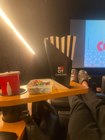 An indoor medium shot captures a wooden tray table in a cinema, likely in Kfar Saba, Israel, with various items on it. A red cup with a white bottle graphic and a straw is placed in a circular holder. Next to it, a clear plastic container holds a collection of colorful candy. To the right, a black popcorn box with white stripes and a logo reading "HOT CINEMA" is prominent. In the background, a cinema screen displays a blue graphic with text. A pair of grey Crocs, adorned with charms, are worn by a person whose legs, clad in black pants, are extended. A black bag is partially visible underneath the table. The scene is illuminated by a bright, diagonal light source on the left, casting shadows and creating an ambiance typical of a movie theater.