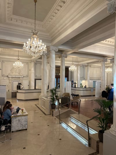 The image displays the grand lobby of the Beau-Rivage Palace hotel in Lausanne, Switzerland. The expansive interior features a high ceiling with intricate white classical molding and cornices, supported by multiple fluted white marble-effect Corinthian columns and thick rectangular beams. Numerous large, illuminated multi-tiered crystal chandeliers are suspended from the ceiling.

The foreground floor is composed of polished light-colored marble tiles with dark square inlays. In the mid-ground, a short, wide set of steps or a low ramp with integrated strip lighting and dark metal handrails leads to a slightly raised section with a darker, possibly wooden, floor.

In the foreground-left, two women are seated at a temporary, rectangular marble-topped desk. One woman, with dark hair, wearing a light blue shirt, is observing a silver laptop. A vase containing small orange flowers is also on this desk. A white face mask lies on the floor near their chairs. Another individual is partially visible to the far left.

In the background, a long, curving main reception desk with a white, textured, diamond-patterned front is visible to the left of center, with a staff member present behind it. A similar, smaller reception desk with a staff member is positioned further to the right. Various lounge areas with upholstered armchairs in cream and gray, accompanied by low tables, are situated in the mid-ground right. Potted green plants are placed throughout the lobby. A man in a dark suit jacket is partially visible from the back in the mid-ground right.