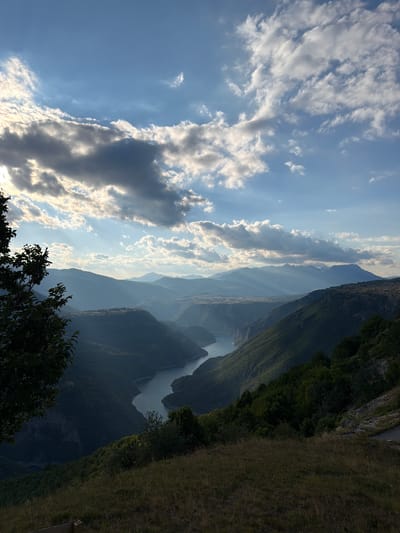 The image captures a panoramic view of a river valley at sunset, with the location identified as being near Dubljevići, Montenegro. The foreground features dry grass and the silhouette of a tree on the left. The middle ground reveals a winding river flowing through a deep canyon, flanked by steep, densely forested mountains. In the background, a series of mountain ranges recede into the distance, veiled in a soft, atmospheric haze. The sky is a dramatic composition of blue, punctuated by scattered, luminous clouds. Sunbeams pierce through the clouds, casting distinct rays of light across the mountainous landscape, creating a striking interplay of light and shadow. The scene conveys a sense of natural grandeur and tranquility.